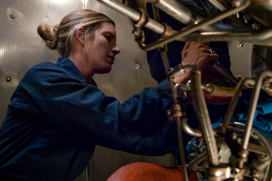 A sailor manipulates a metal pipe in a dark industrial-type room.
