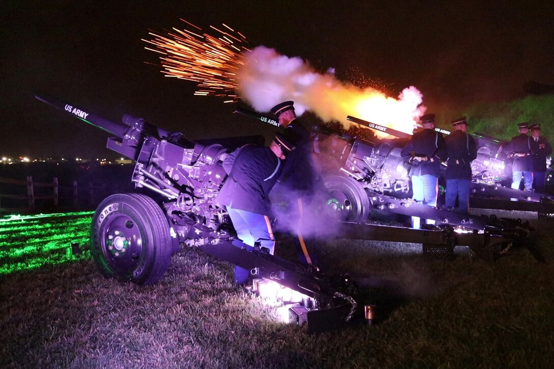 Soldiers fire cannons at a park against a night sky lit up by the blast and by purple and green light.