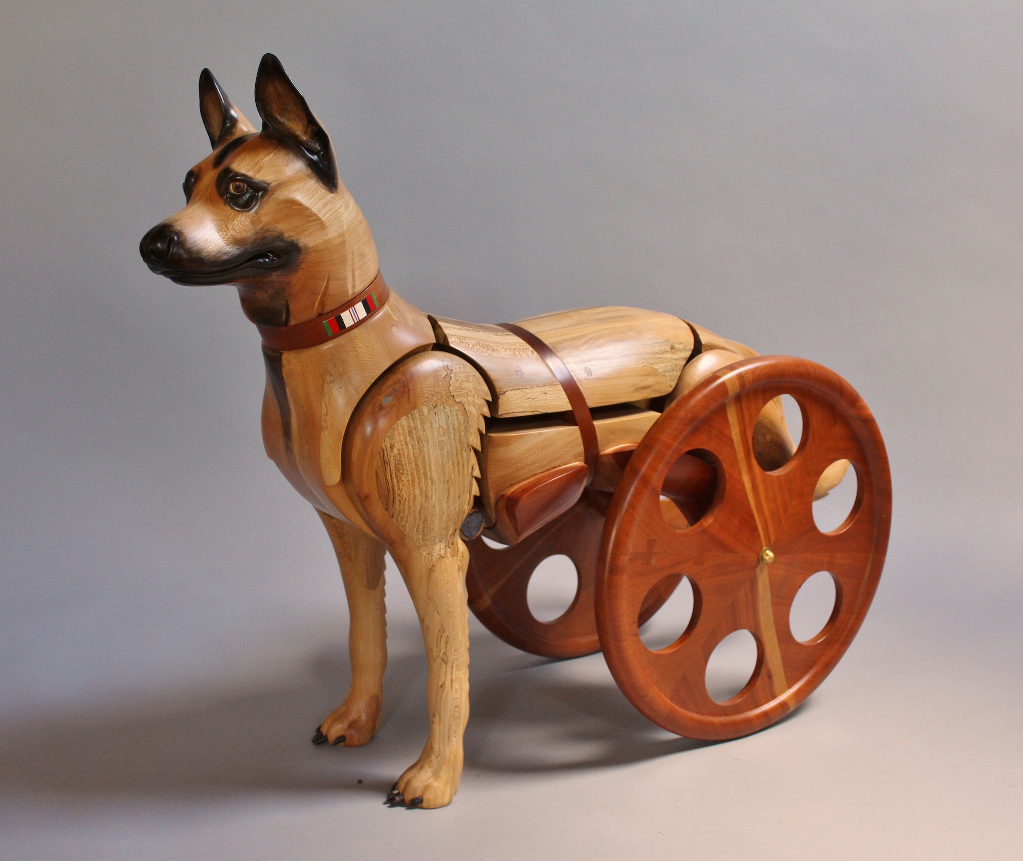 Wooden sculpture depicting a dog with its hind legs amputated and using a wheelchair.