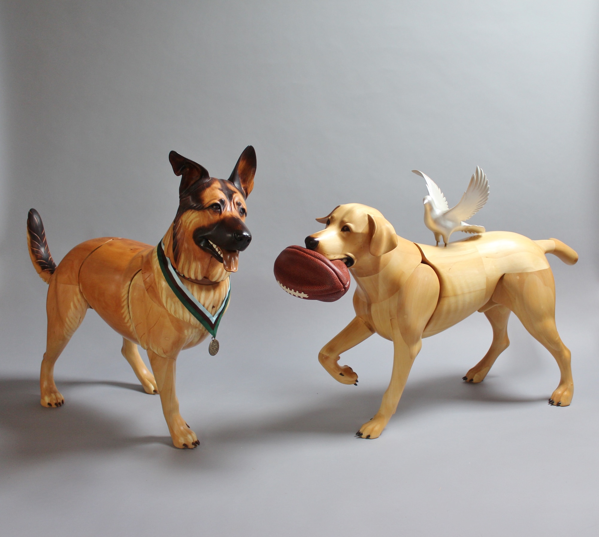 Picture of wooden sculpture dogs. The dog on the left, Lucca, has his front leg amputated and is wearing a medal around her neck. The dog on the right, Cooper, has a deflated football in her mouth and a white dove on her back.