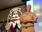 Man in tan Navy uniform talks in front of flags.