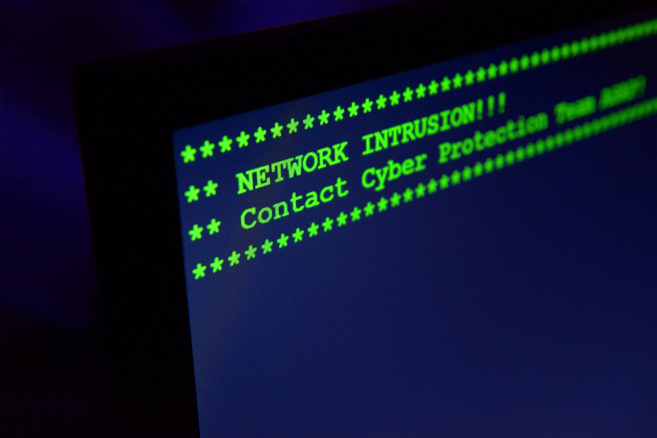 In a dark room, the words "NETWORK INTRUSION!!! Contact Cyber Protection Team ASAP" flash on a computer screen.