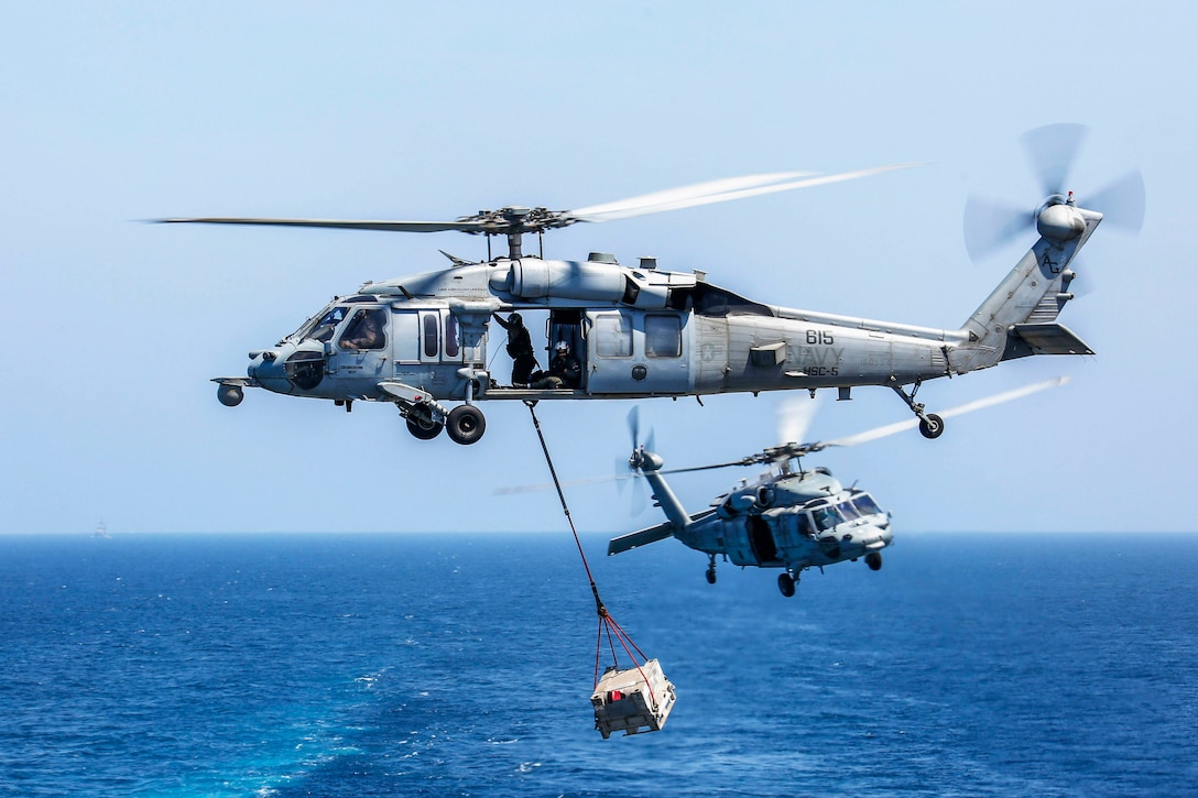 Two helicopters fly; one carrying cargo underneath.