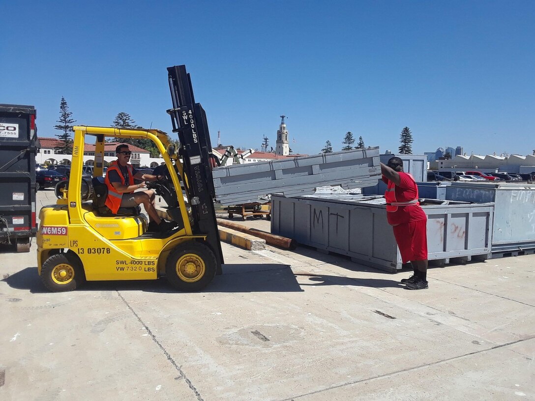 The person on the left uses a forklift to pick up scrap with a person tot he right guiding the scrap