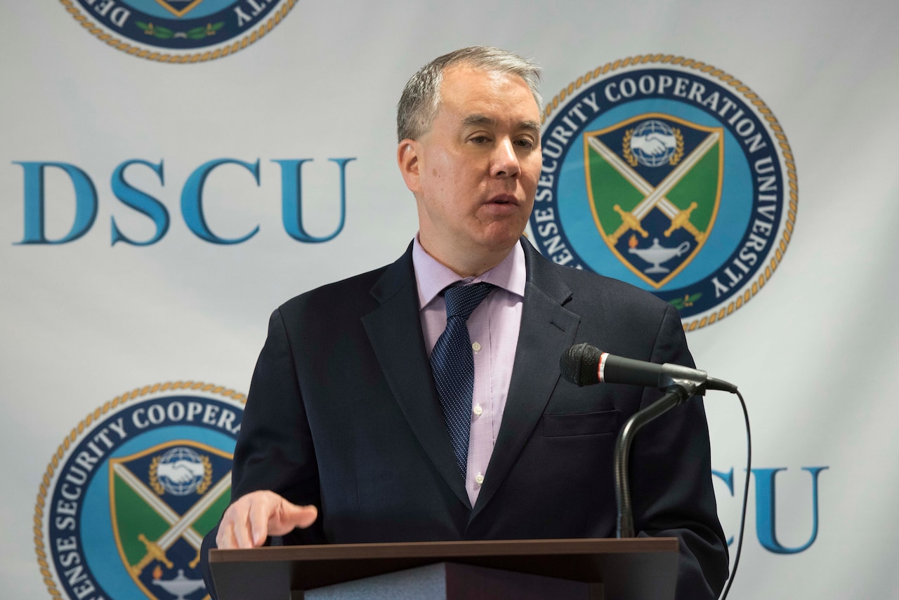 A man speaks at a lectern. Behind him, the letters “DSCU” and a logo are on a backdrop.