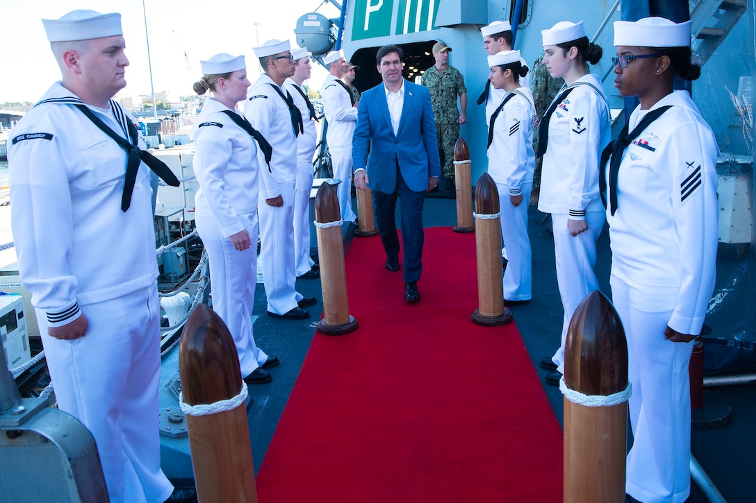 Defense Secretary Dr. Mark T. Esper walks along a red carpet with a row of sailors on each side.