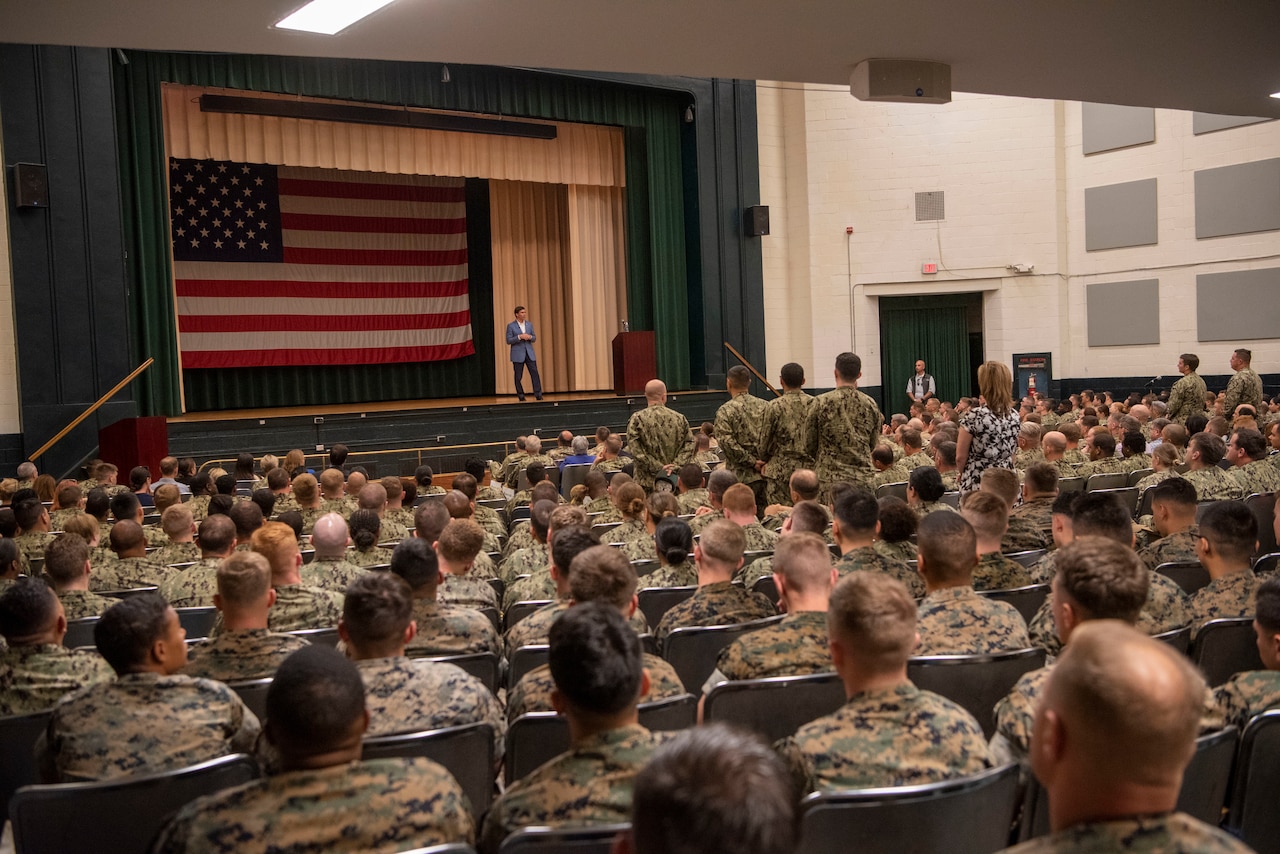 Man wearing suit stands on a stage in front of a large U.S. flag. He is speaking to an audience of men and women -- most dressed in military fatigues.