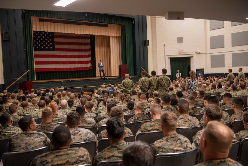 Man wearing suit stands on a stage in front of a large U.S. flag. He is speaking to an audience of men and women -- most dressed in military fatigues.