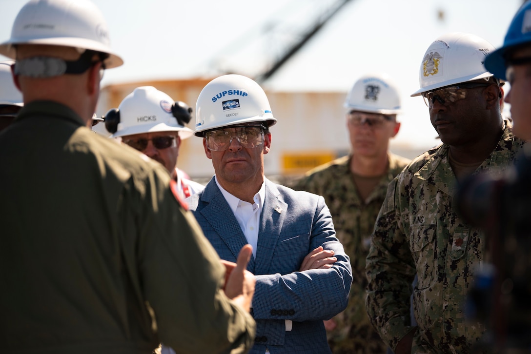 Man wearing hard hat and safety glasses listens to a man in a green uniform and hard hat, whose back is to the camera.
