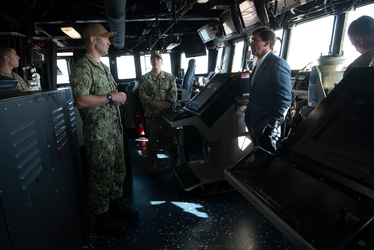 Aboard a ship, a man in a suit talks to a man dressed in fatigues as three other men look on.