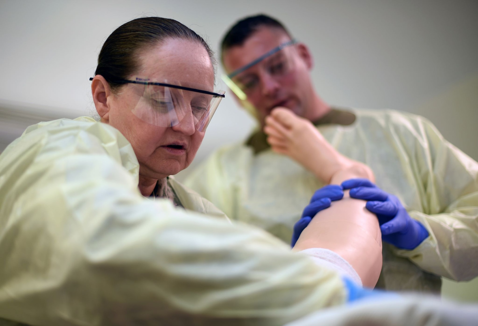 One female and one male medical professional with latex gloves wipe gauze along the skin of a training mannequin. The training mannequin has rubber covering on it simulating a burn.