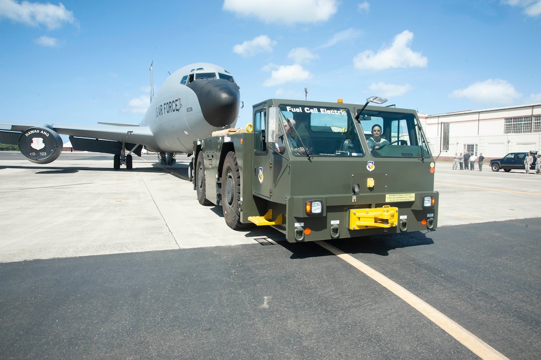 A green flatbed tow vehicle with “Fuel Cell Electric” written on the windshield pulls a large military transport jet.