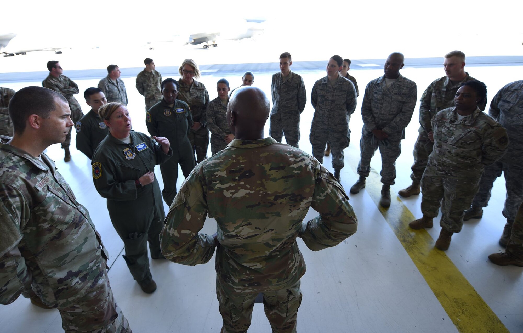 Chief Wright is surrounded by Airmen in a brightly lit aircraft hangar. He's speaking to them.