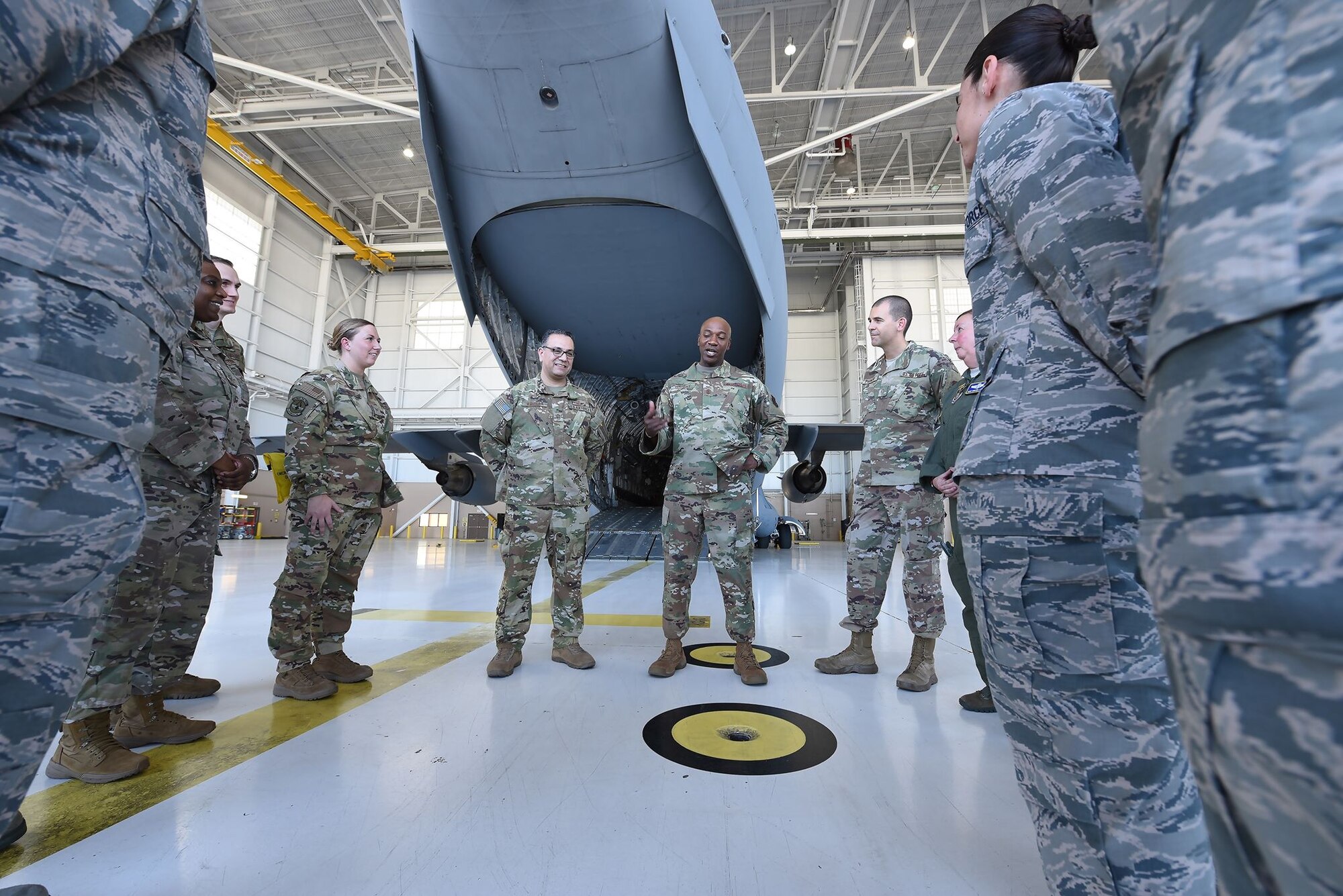 Chief Wright stands in front of the back opening of a C-5M Super Galaxy in a brightly-lit aircraft hangar. Airmen surround him. They're listening to what he's saying intently.