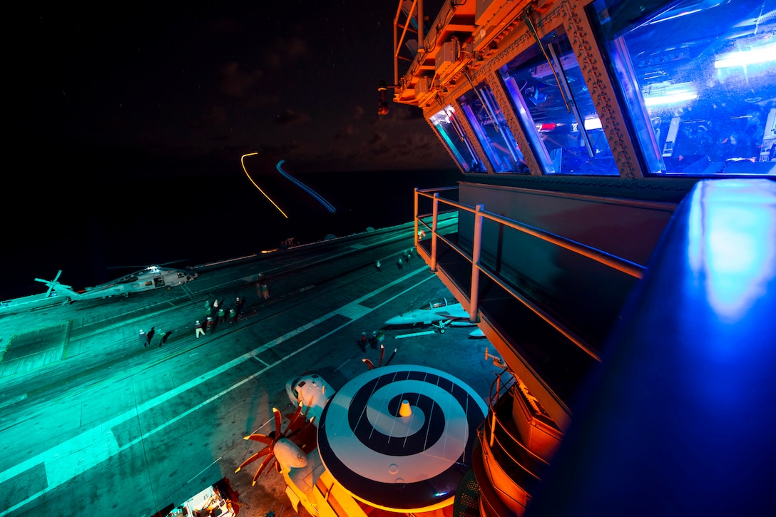 A military helicopter prepares to take off from a brightly lit aircraft carrier at night.