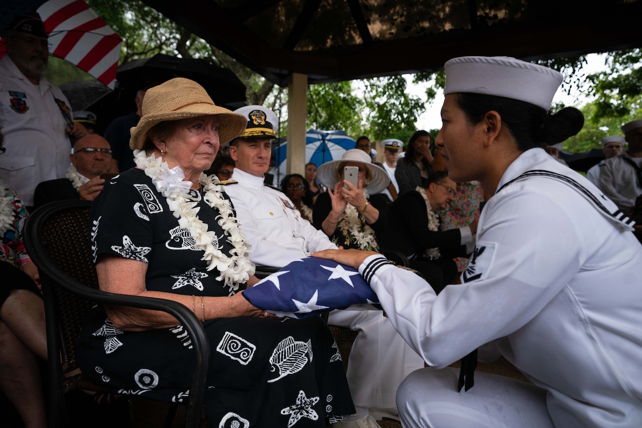 A sailor places a flag in the hands of a seated woman  during a funeral.