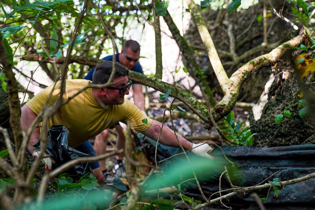 A sailor reaches for trash in a wooded area as another person works behind him.