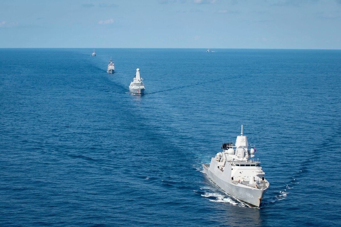 A group of ships move in formation in a straight line.