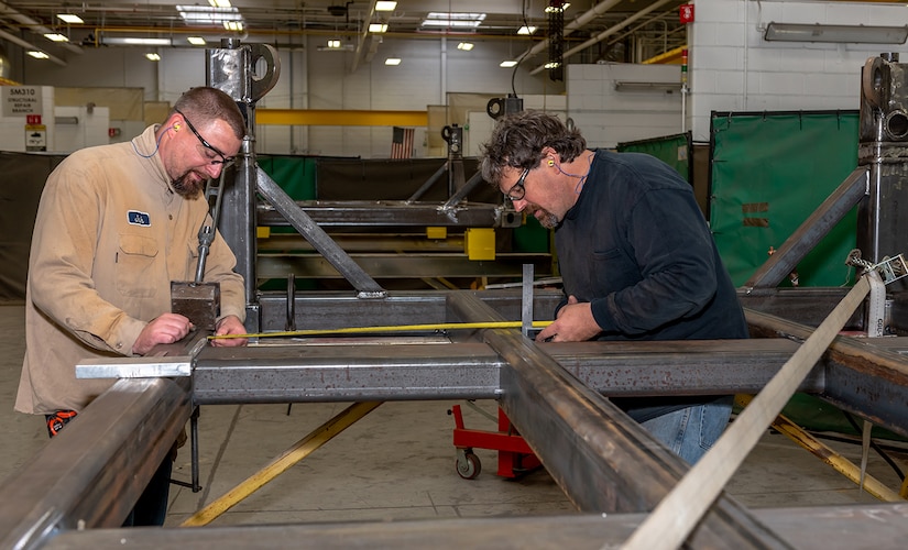 Photo of Welder and Welding Worker measure the PEG pallet frame assembly to mark the location of the cross beam.