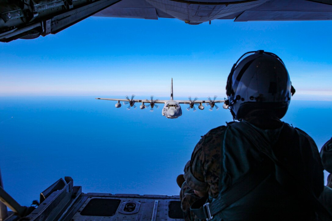 A service member looks out the back of a military plane at another large military aircraft flying above the ocean.