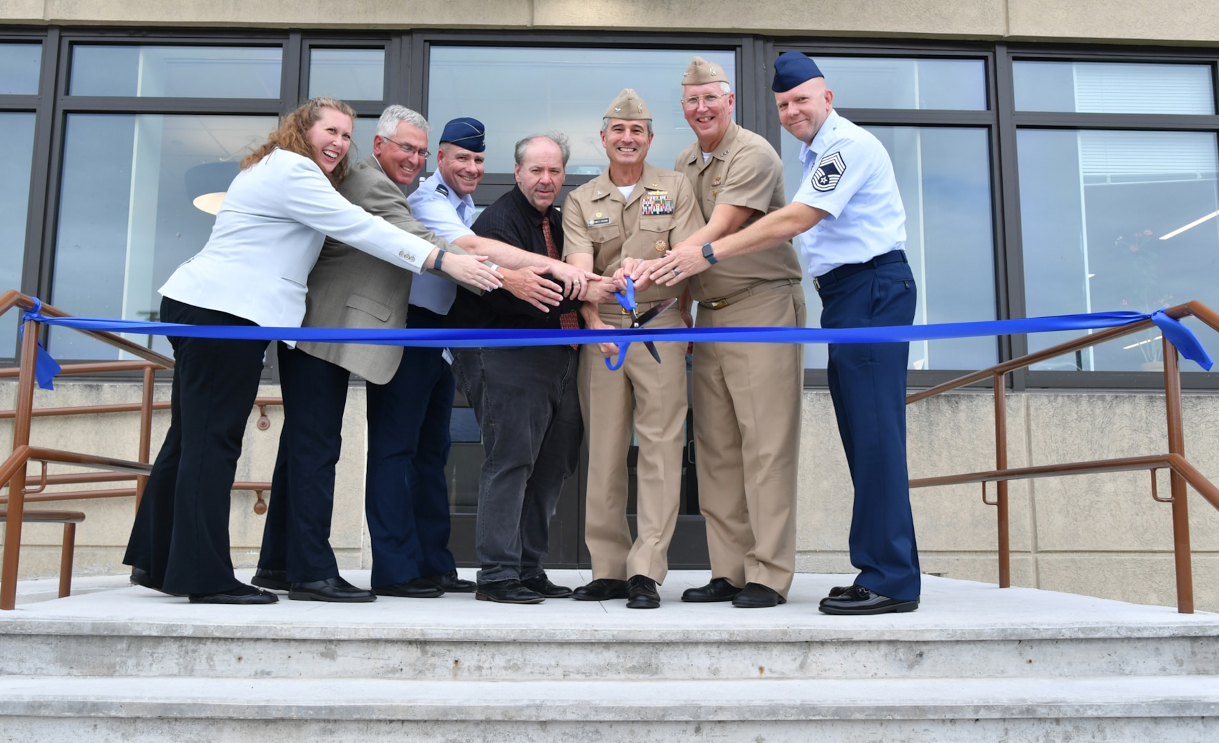 Group shot of military and civilian personnel cutting a blue ribbon in front of a building
