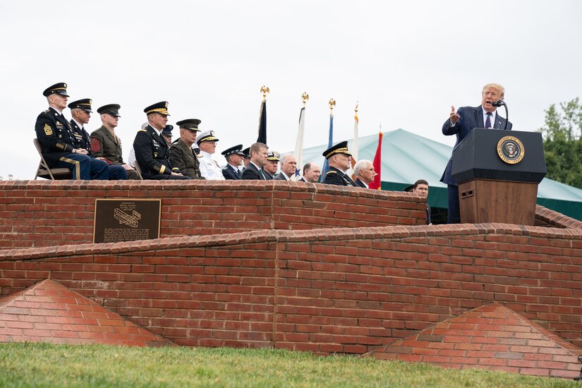 President gestures while speaking at a lectern with military and civilian officials seated nearby.
