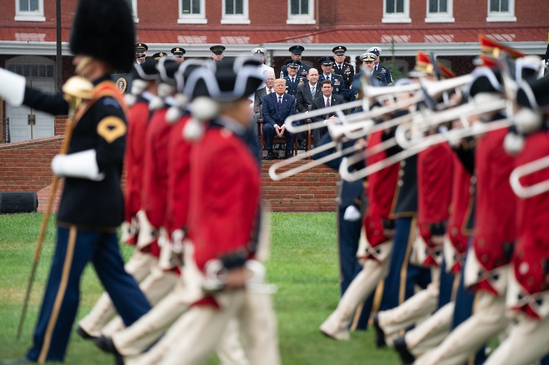 Military musicians march on a field as senior leaders watch.