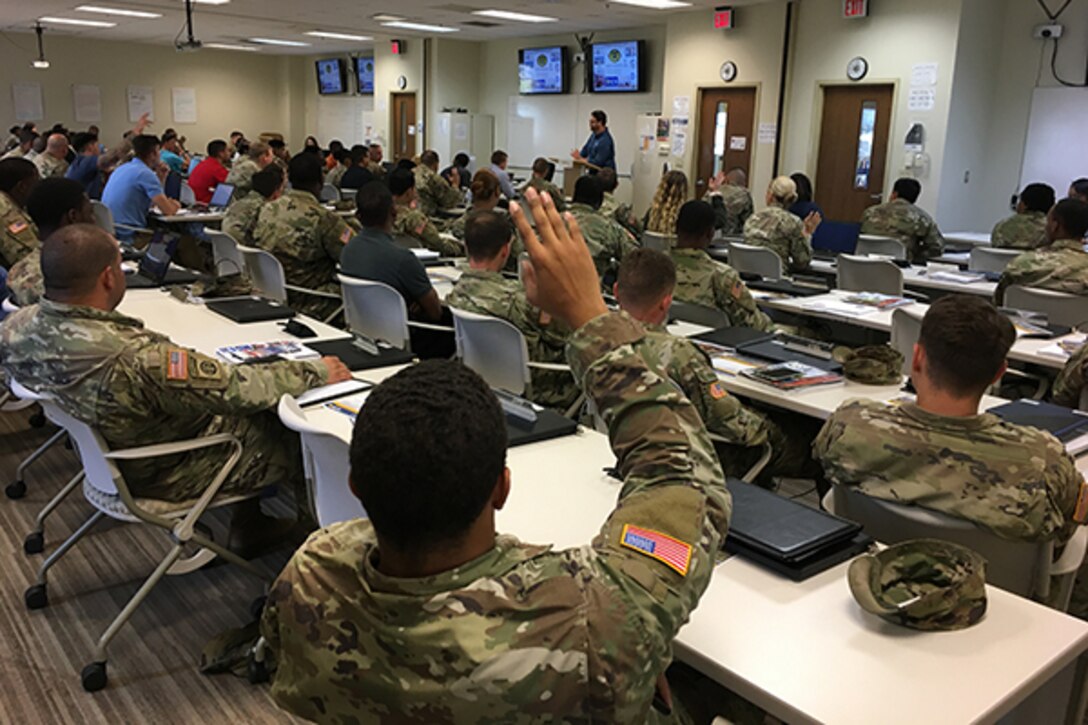 Man speaks to soldiers in a classroom setting as soldier in foreground raises hand to ask a question.