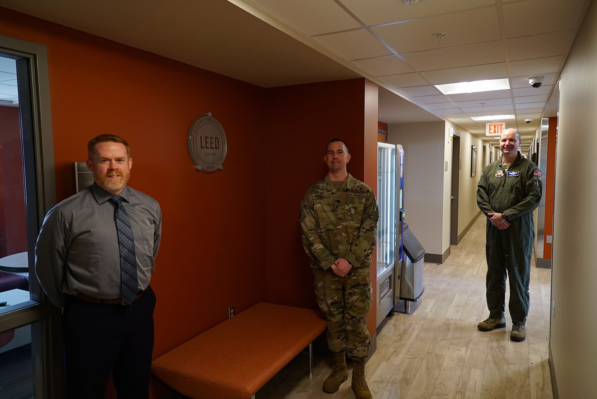 Three men stand in the first-floor hallway of Gray Hall Dorm near a clouded glass placard that reads "LEED," hanging on a reddish-brown wall.