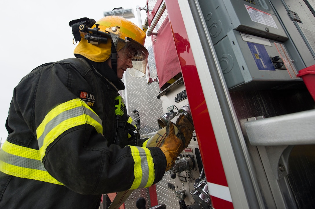An airman in an fireman's suit connects a hose to a firetruck.