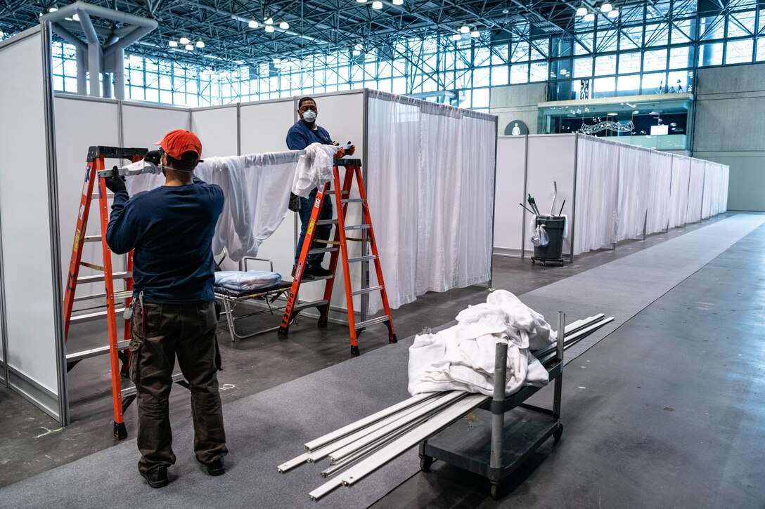 Two men set up partitions and curtains on a large convention center floor.