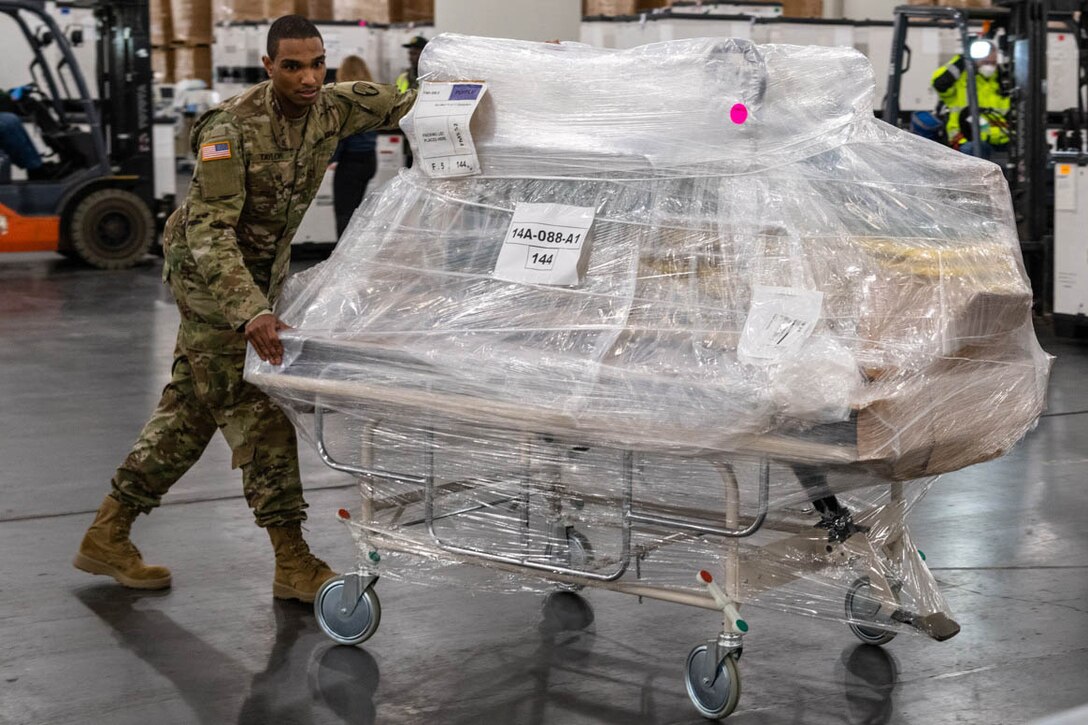 A service member moves a mobile hospital bed that is also loaded with gear.