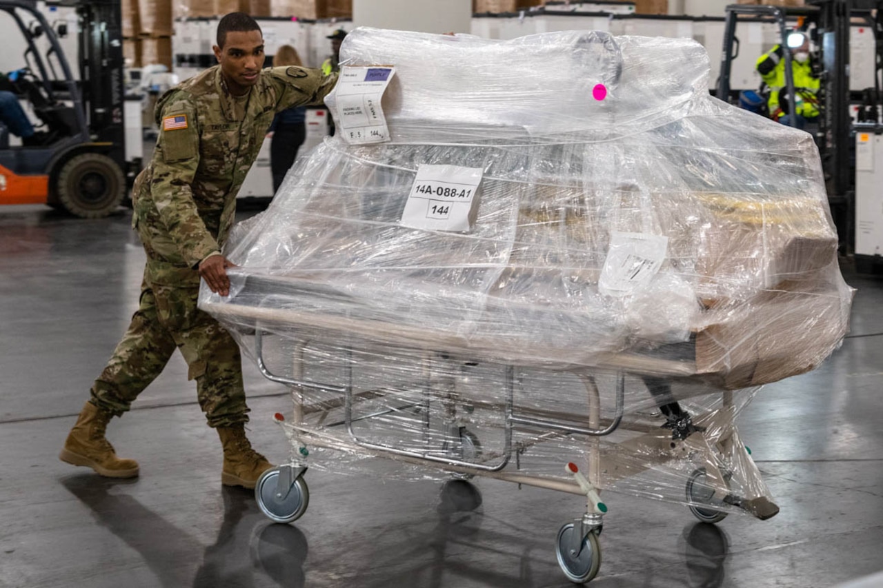 A service member moves a mobile hospital bed that is also loaded with gear.