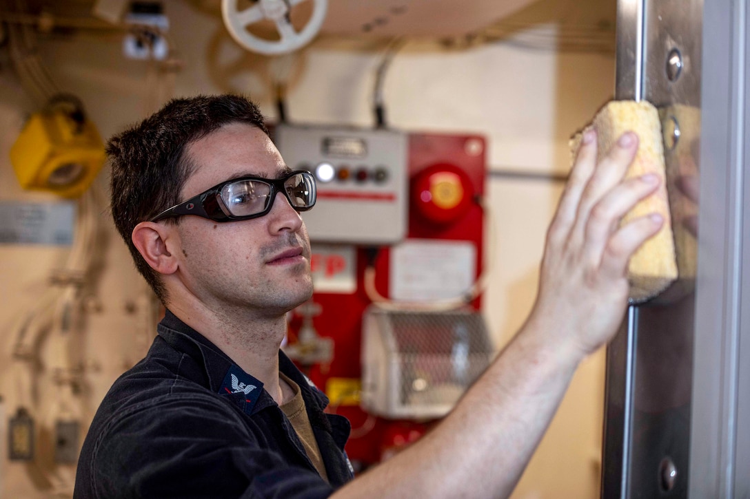 A sailor runs a sponge over a metal surface on a ship.