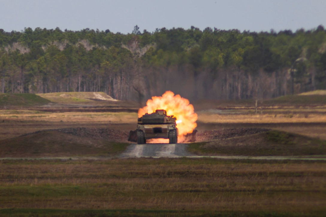 A tank fires a large blast in the middle of a field; a tree line seen in the background.