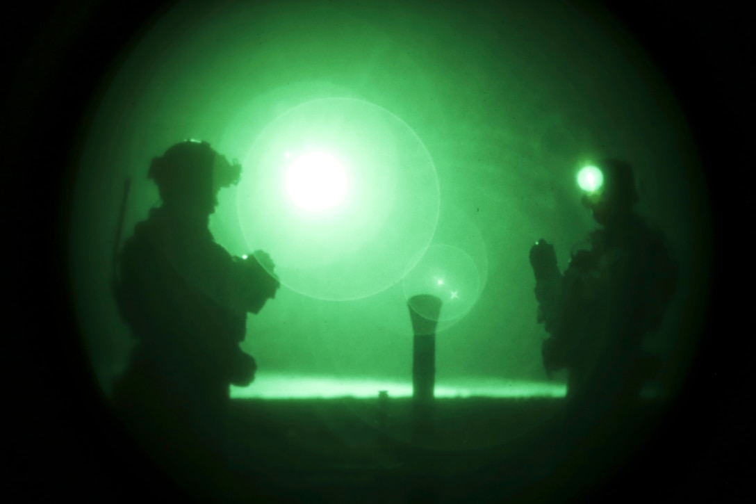 Two soldiers, shown in silhouette and illuminated by green-light, stand by a mortar system on the ground.