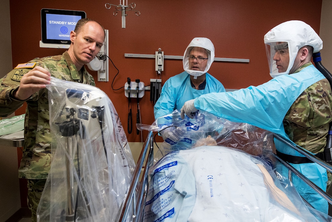 Two military doctors in uniform remove plastic sheeting from medical equipment as they discuss procedures with a third doctor dressed in civilian clothes.