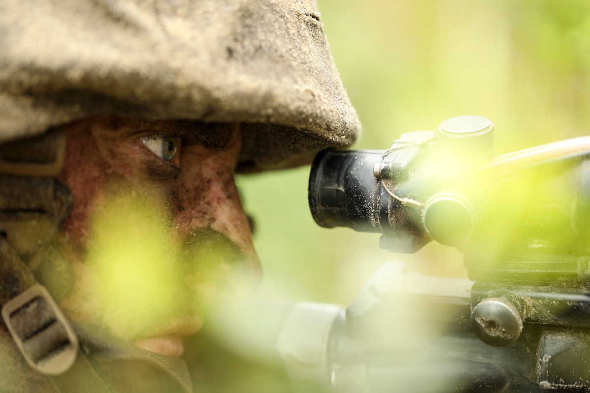 A Marine Corps recruit, shown in a closeup profile, looks through a weapon scope while obscured by foliage.