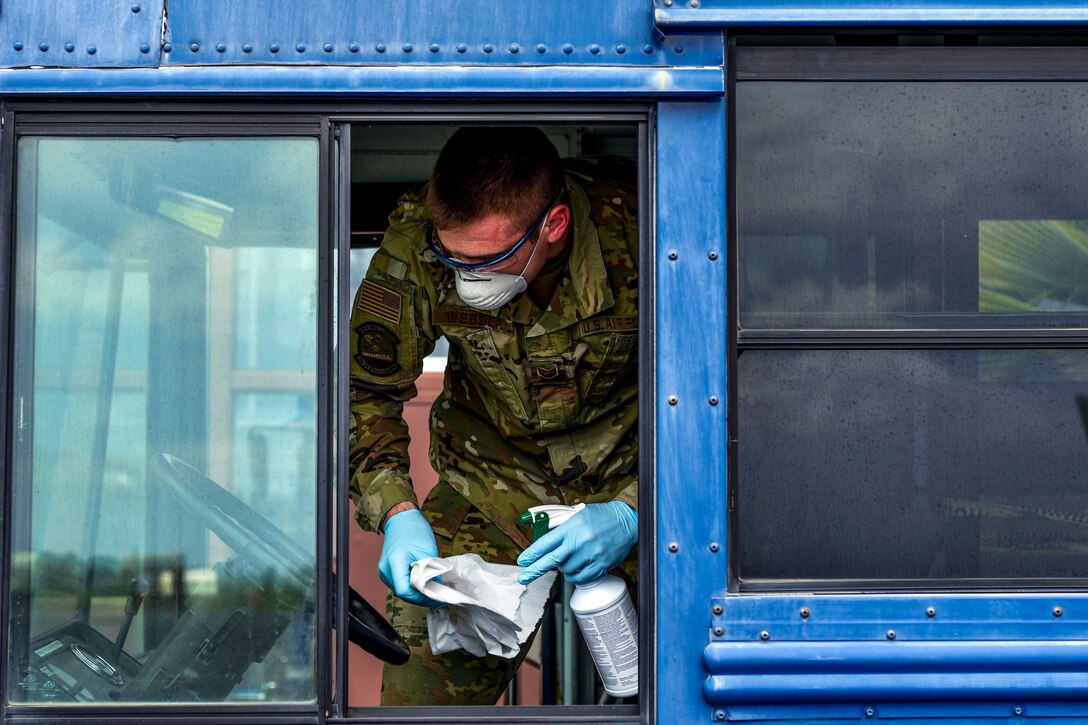 An airman wearing a mask and gloves cleans on a bus.