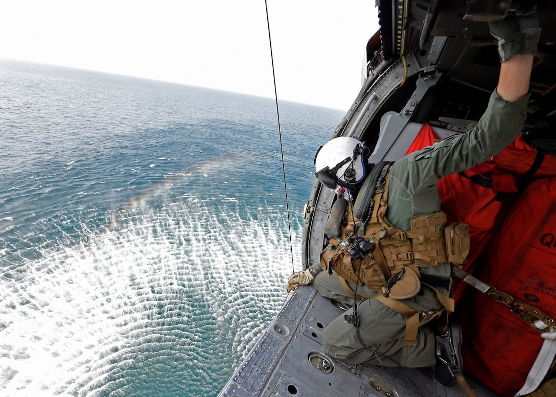 A sailor looks over the side of an open helicopter flying over water.