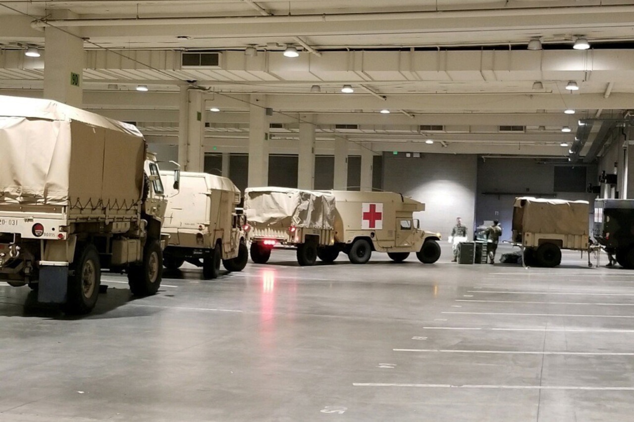 A line of military vehicles, one with a red cross on its side, move through a large garage.