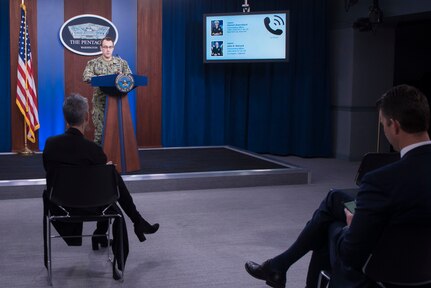 A man in uniform stands at a podium in front of a Pentagon sign and next to a monitor showing the names and pictures of the two captains calling into the briefing.