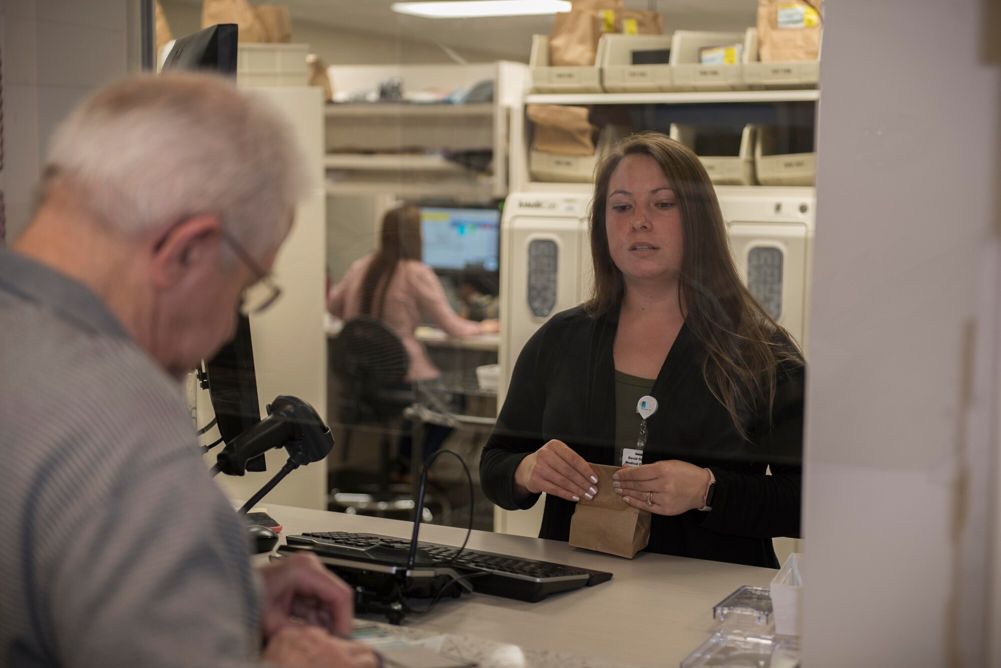 Bria McCarthy, 60th Medical Group pharmacy technician, assists a pharmacy customer while avoiding direct contact March 30, 2020, at Travis Air Force Base, California. Travis AFB pharmacies implemented multiple procedures to combat COVID-19, including placing protective plastic barriers between pharmacy technicians and customers. (U.S. Air Force photo by Airman 1st Class Cameron Otte)