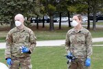 Two soldiers wearing masks and blue gloves stand with medical screening equipment in their hands.