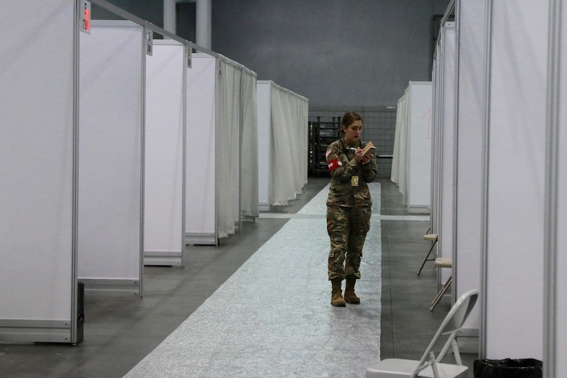 A soldier writes on a notepad as she walks through a corridor of empty makeshift hospital rooms.
