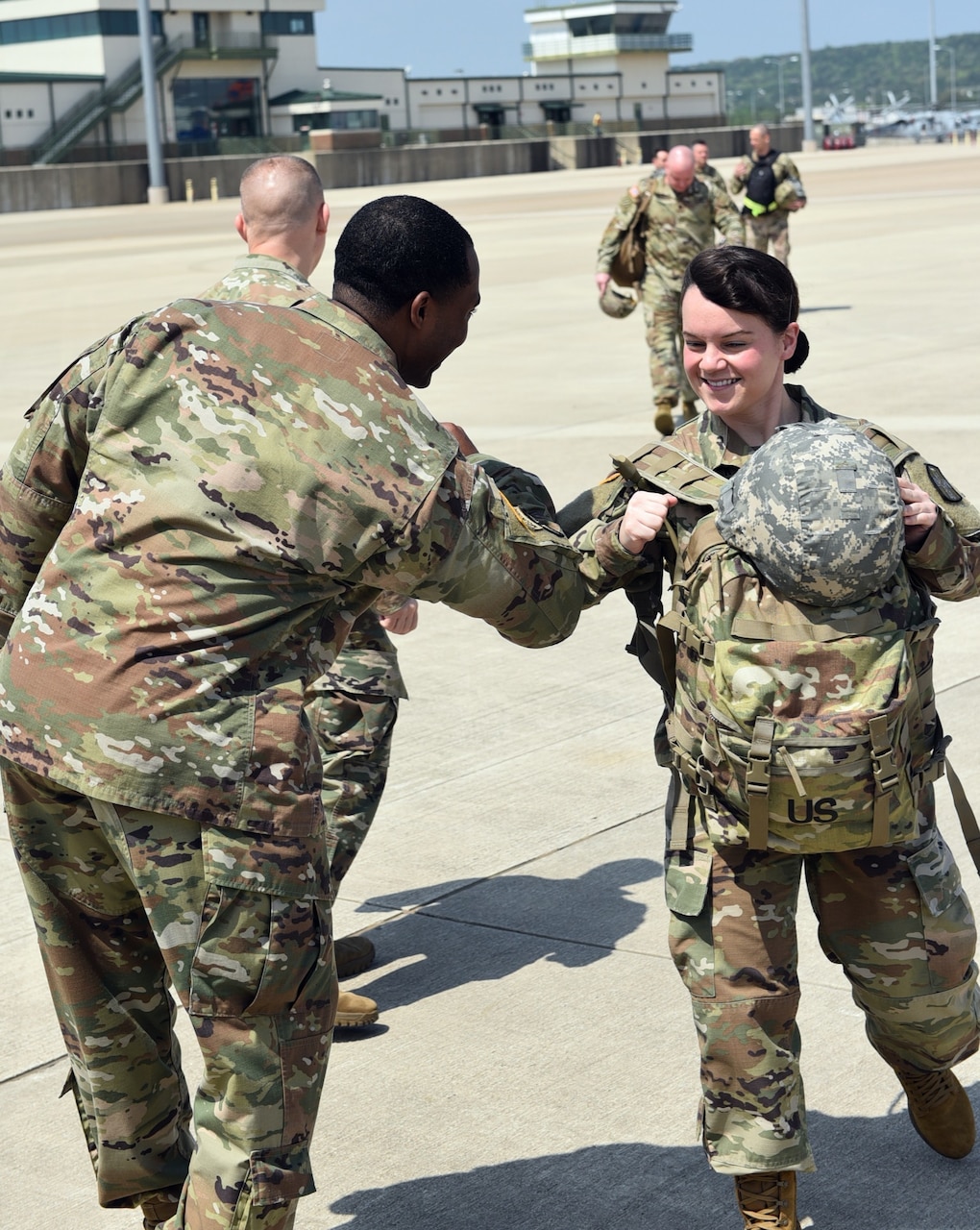 A supervising soldier elbow-bumps a female soldier carrying a heavy pack across tarmac.