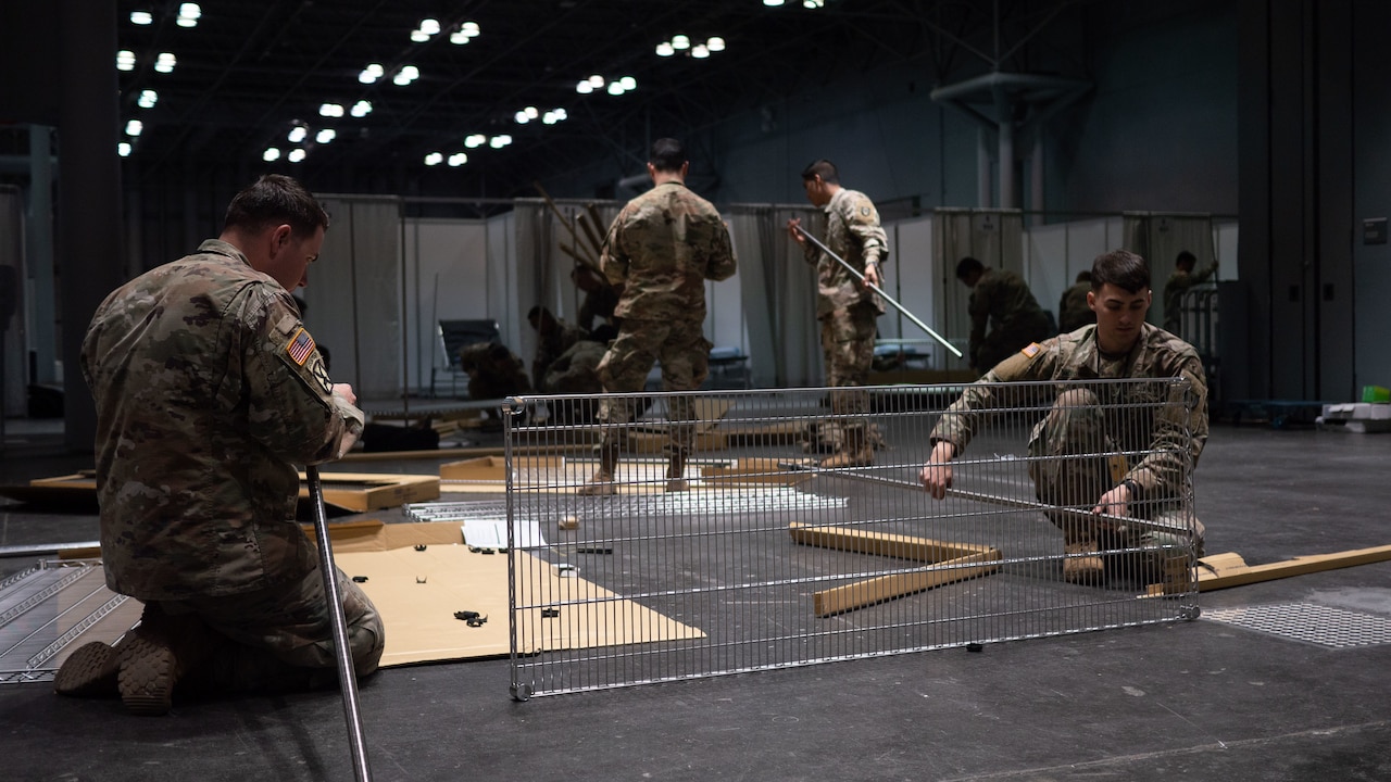 Two soldiers kneel down while putting together temporary shelving inside a large room. Others do similar tasks in the background.