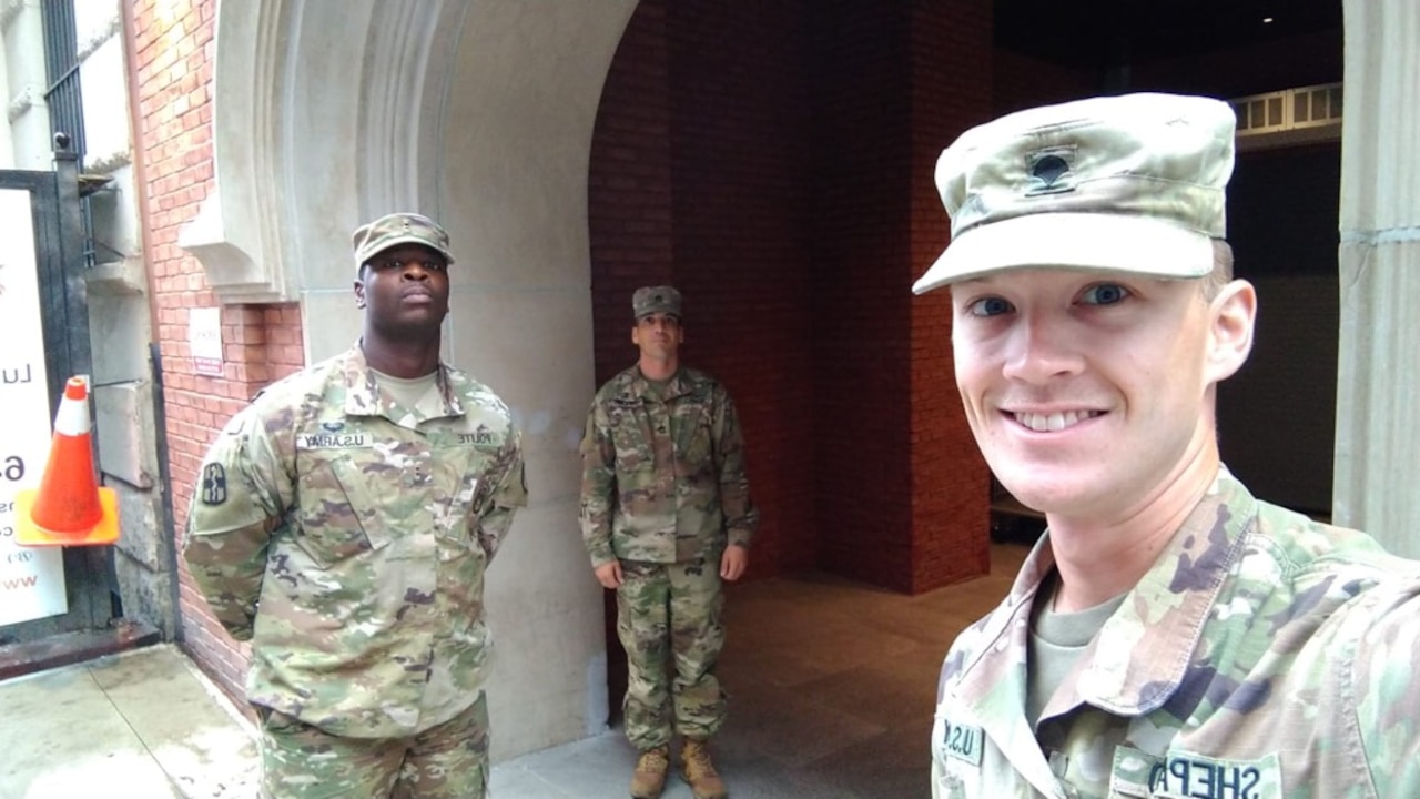 A soldier takes a selfie, while two other soldiers pose far apart in the background.