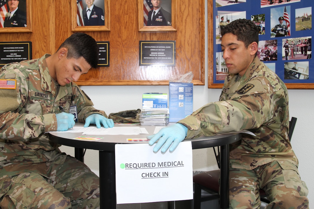 Two soldiers wearing plastic gloves sit at a table with a medical check-in sign hanging from its edge.