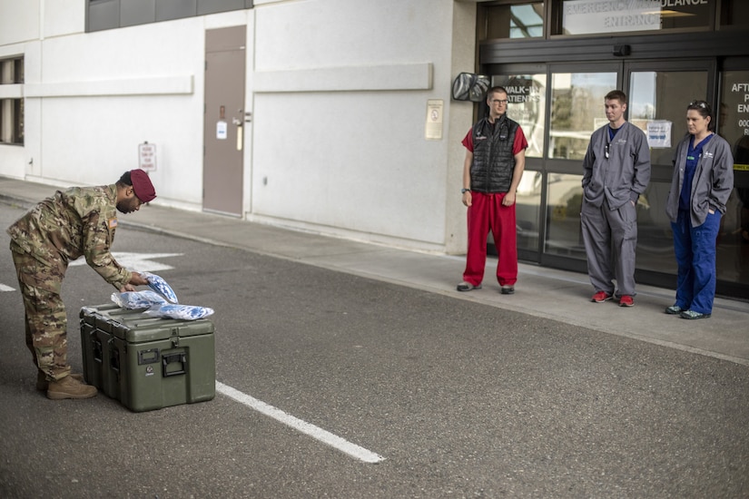 A soldier places bags of masks on a hardened container as medical personnel look on.